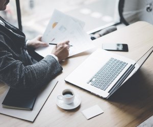 Adult elegant banking finance analyst working at sunny office on laptop while sitting at wooden table.Businessman analyze stock reports.Paper documents holding on hands.Blurred background,horizontal.