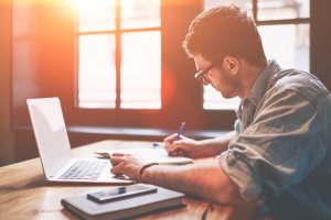 Young millennial male student searching information for homework using laptop and wifi connection indoors, man writing article in textbook for add interesting content on own website or web page