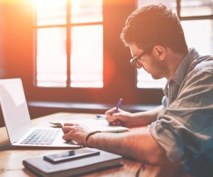 Young millennial male student searching information for homework using laptop and wifi connection indoors, man writing article in textbook for add interesting content on own website or web page